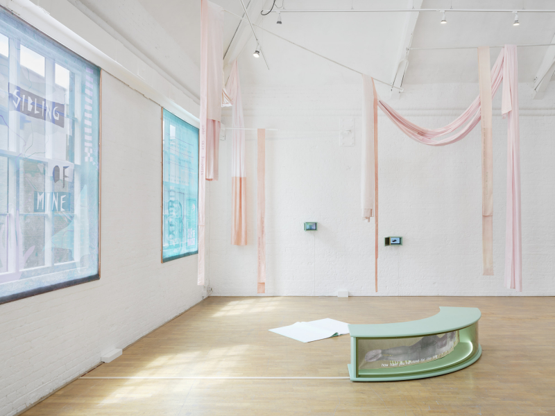 Installation view showing pale pink sheets draped from the ceiling and a pale green curved bench by a window in a bright, white painted room with large windows.